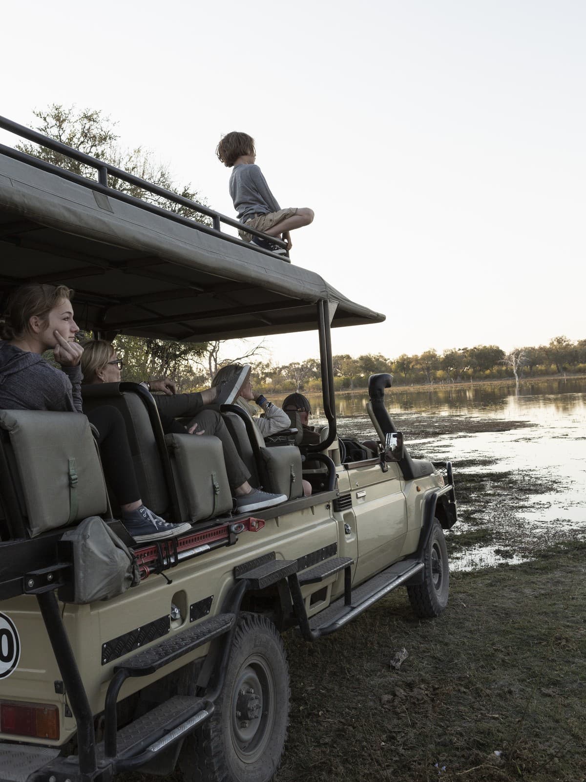 Junge beobachtet vom Dach des Safari-Jeeps ein Wasserloch, Familie sitzt im Fahrzeug