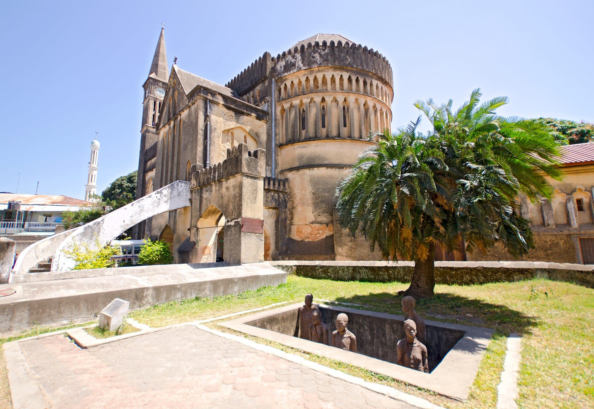 Anglikanische Kathedrale in Stone Town auf dem Gelände des früheren Sklavenmarkts, davor das Denkmal mit Figuren in einer Grube, im Hintergrund ein Minarett