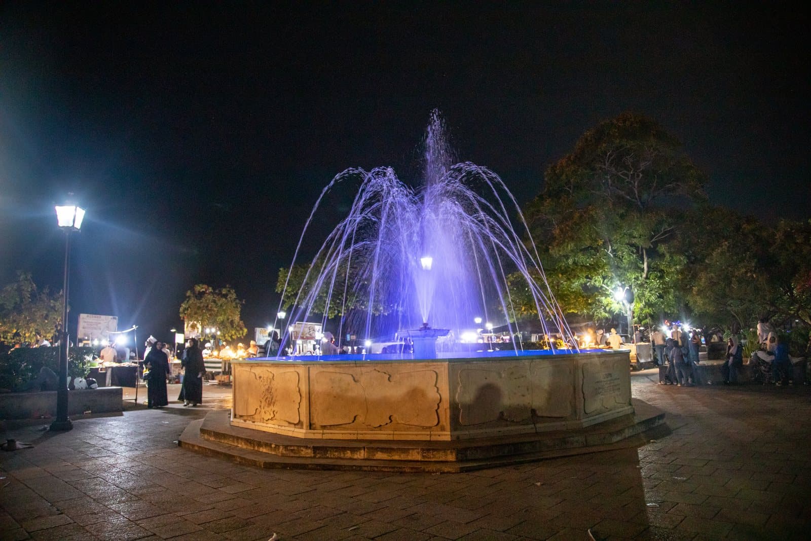Beleuchteter Springbrunnen in den Forodhani-Gärten in Stone Town am Abend, dahinter Marktstände