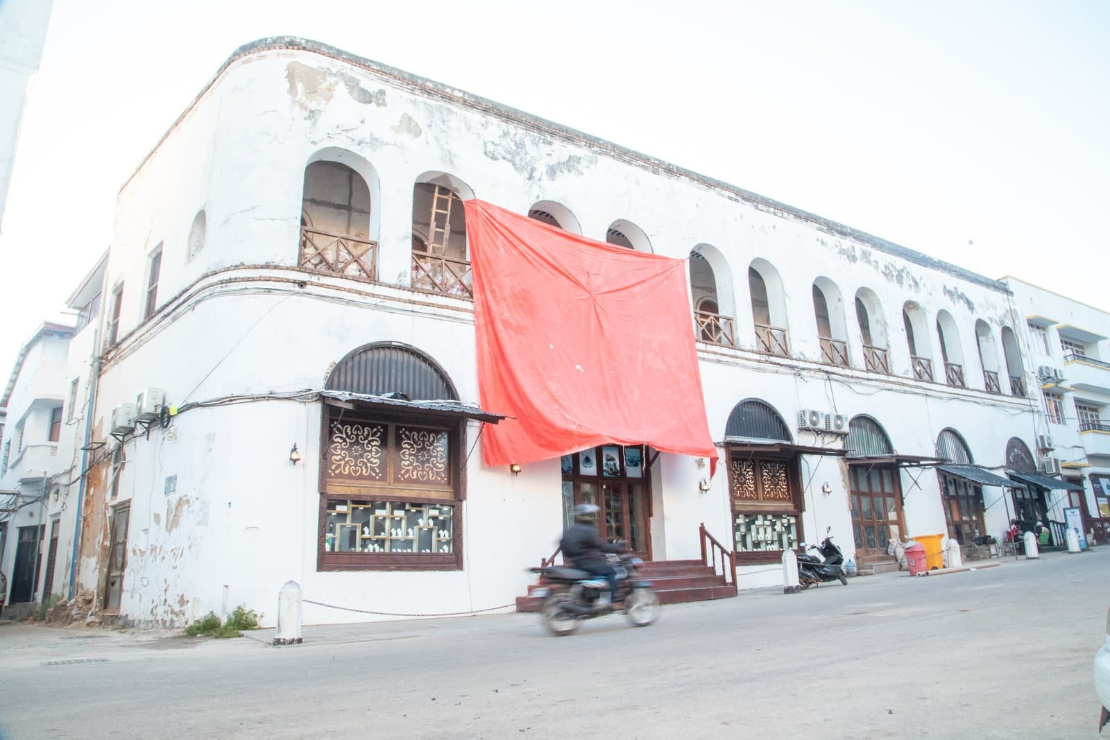 Weiß getünchtes Gebäude mit Bogenfenstern und Holzbalkonen an einer Straße in Stone Town, davor ein Motorrad