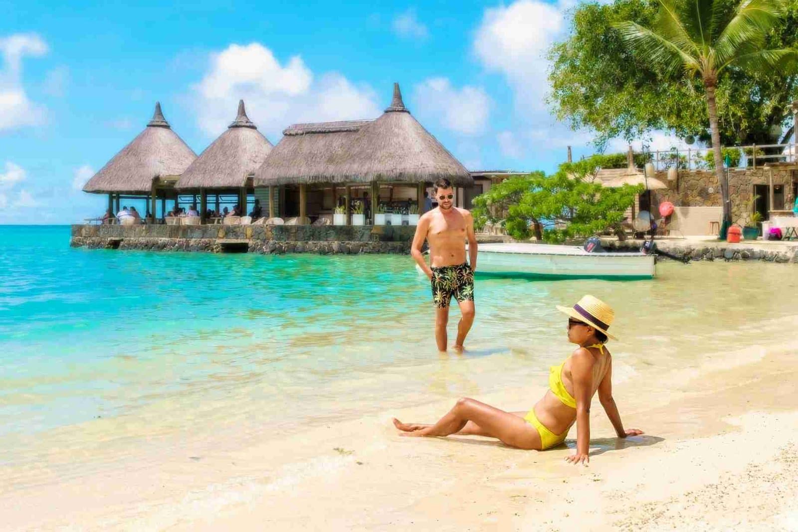 Paar am Strand vor einer Strandbar mit Strohdächern und türkisfarbenem Wasser